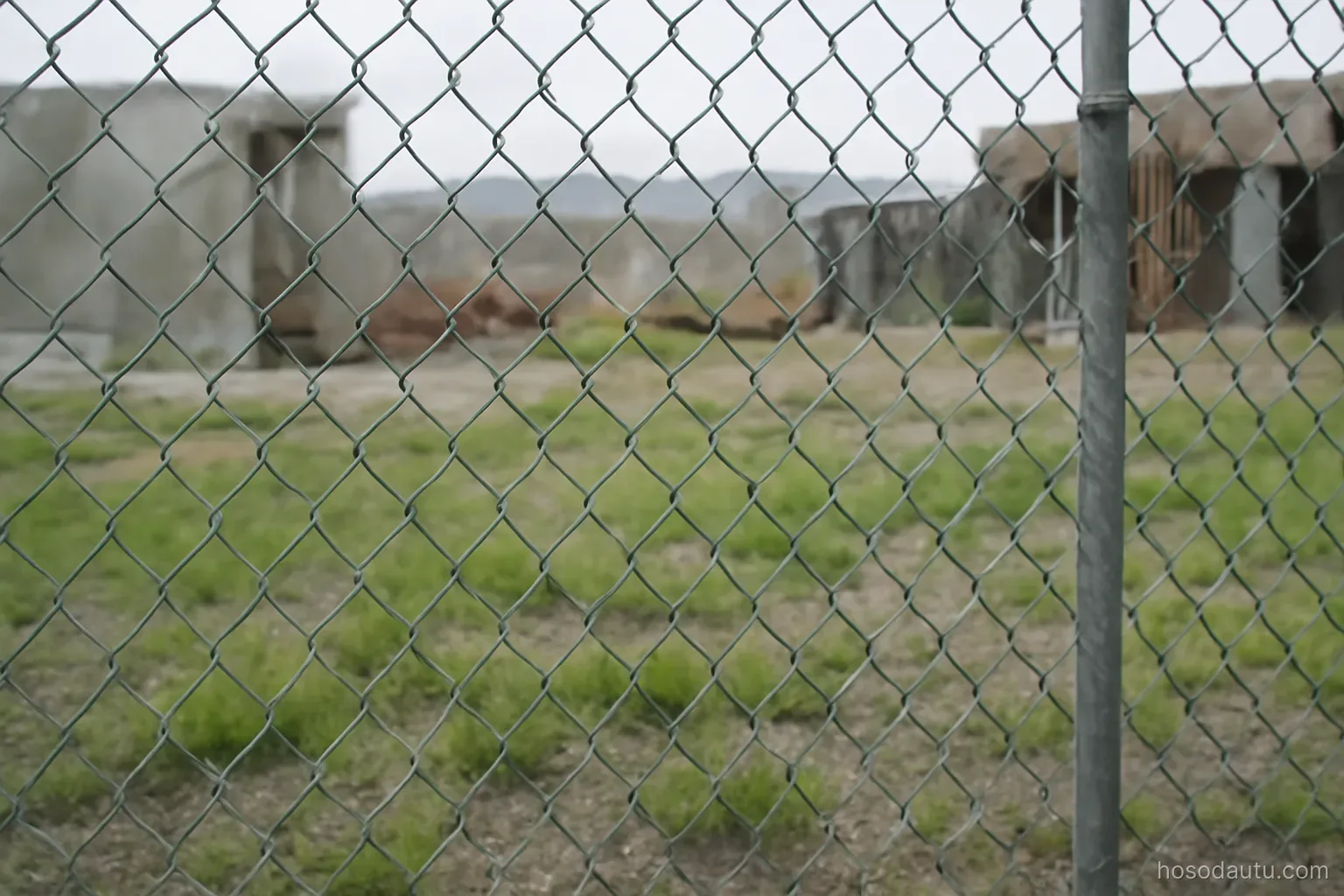 a chain link fence around an idle construction site with grass growing through gravel, grey daylight, no signs
